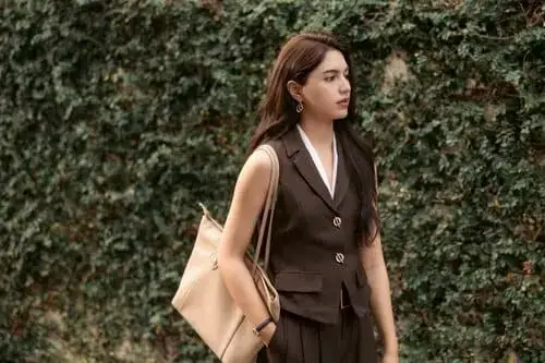 by-your-side-the-series-hinh-anh-9 By Your Side nữ chính mặc vest nâu đeo túi tote đứng trước hàng rào cây xanh